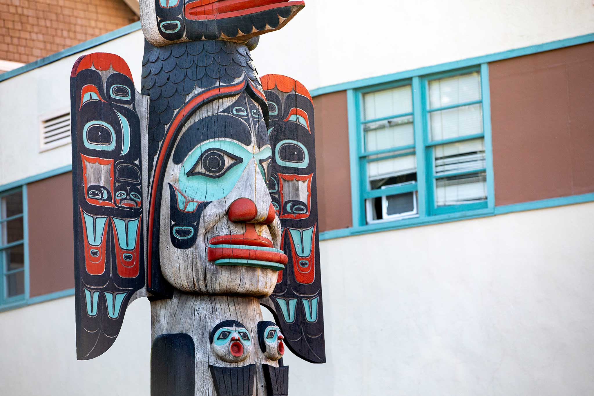Colorful Alaska Native totem pole with expressive faces and symbols in front of a building.