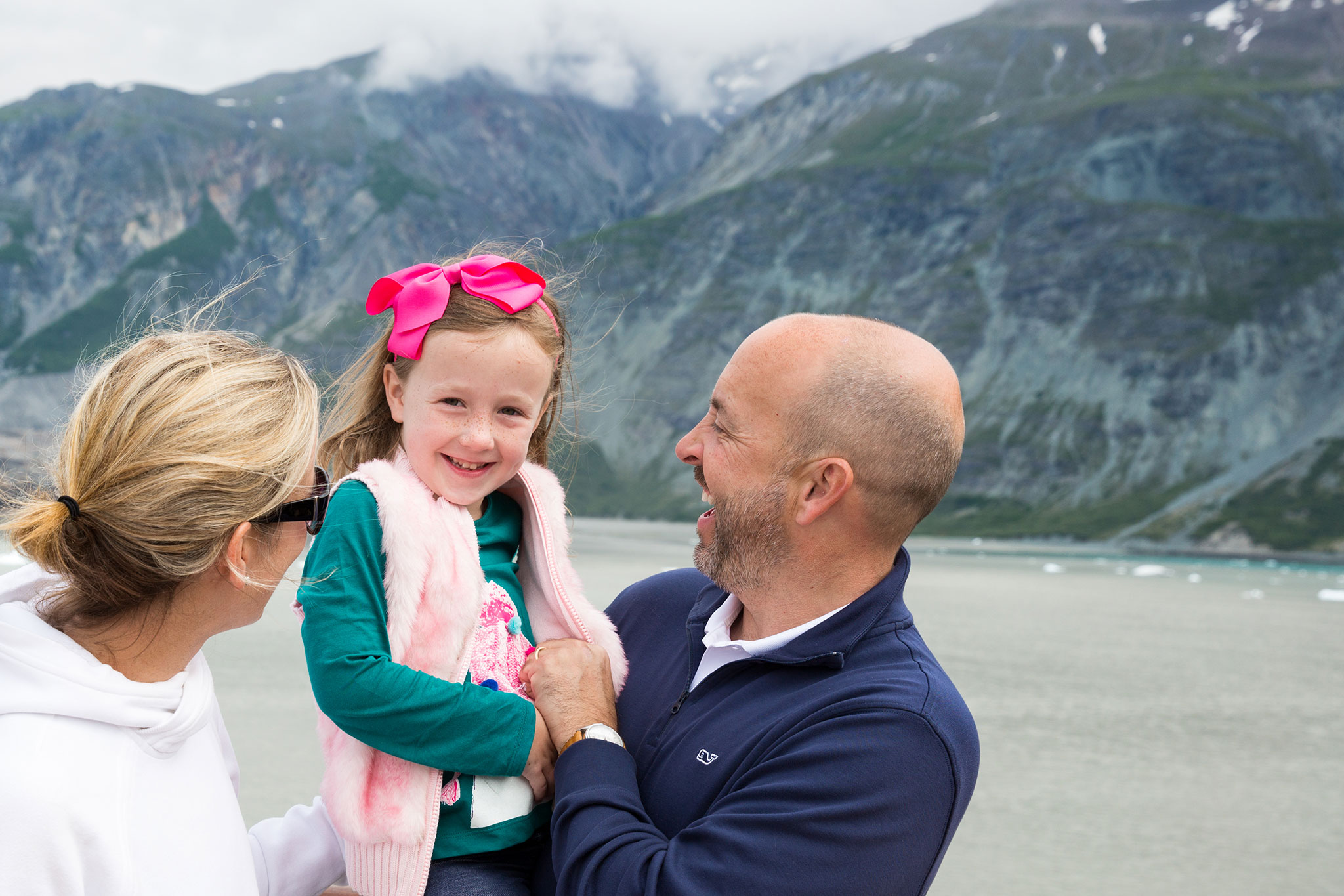 A smiling little girl with a pink bow held by her parents in front of a glacial backdrop.