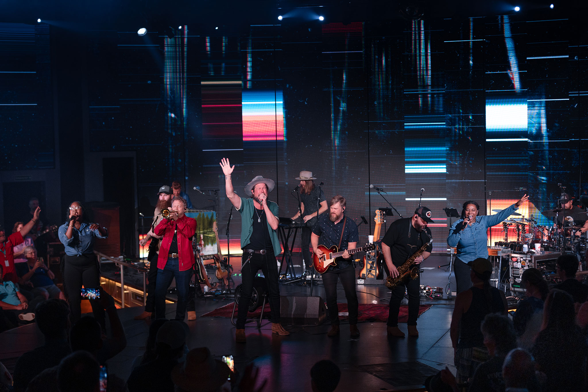 Live concert scene featuring a diverse group of musicians, including brass players and guitarists, with dynamic digital backdrops.