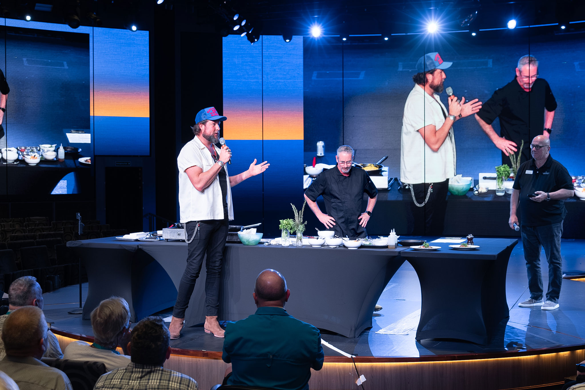Host interacting with a chef during a cooking show in front of an engaged audience, with vivid backdrop screens.