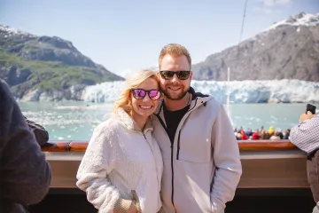 Smiling couple in sunglasses posing in front of a glacier from a cruise ship.