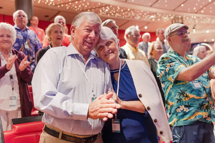 An elderly couple embracing and smiling joyfully at a social event, surrounded by a crowd of applauding attendees.